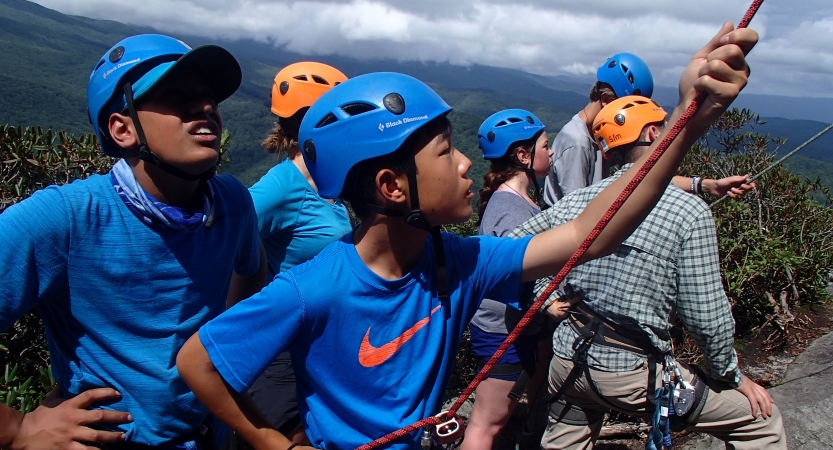 Students wearing helmets and harnesses belay rock climbers, while others spot them. They appear to be at high elevation, above a green forrest. 